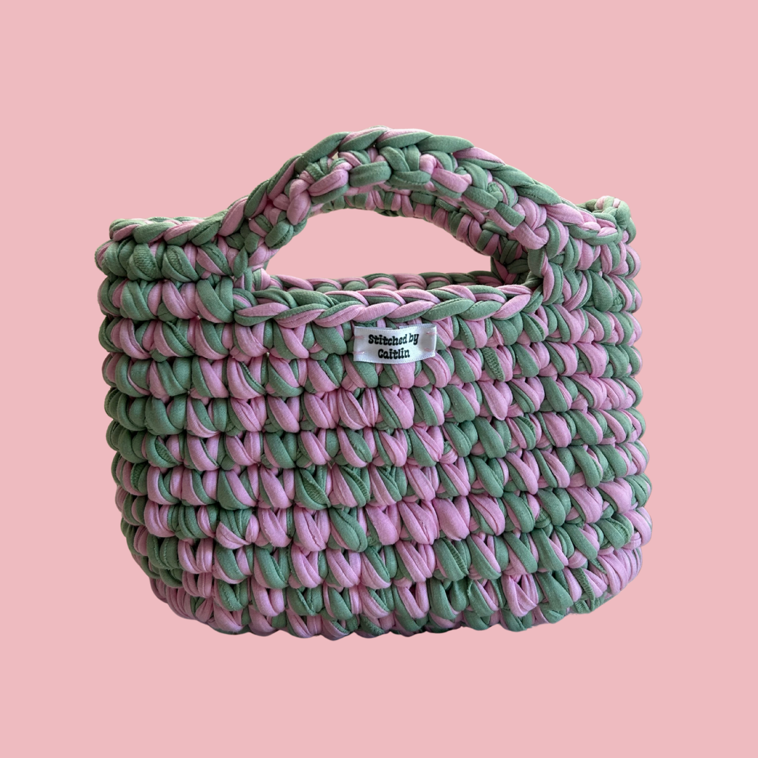 Pink and mint crocheted bag with two small handles at the top and a Stitched by Caitlin label on the front