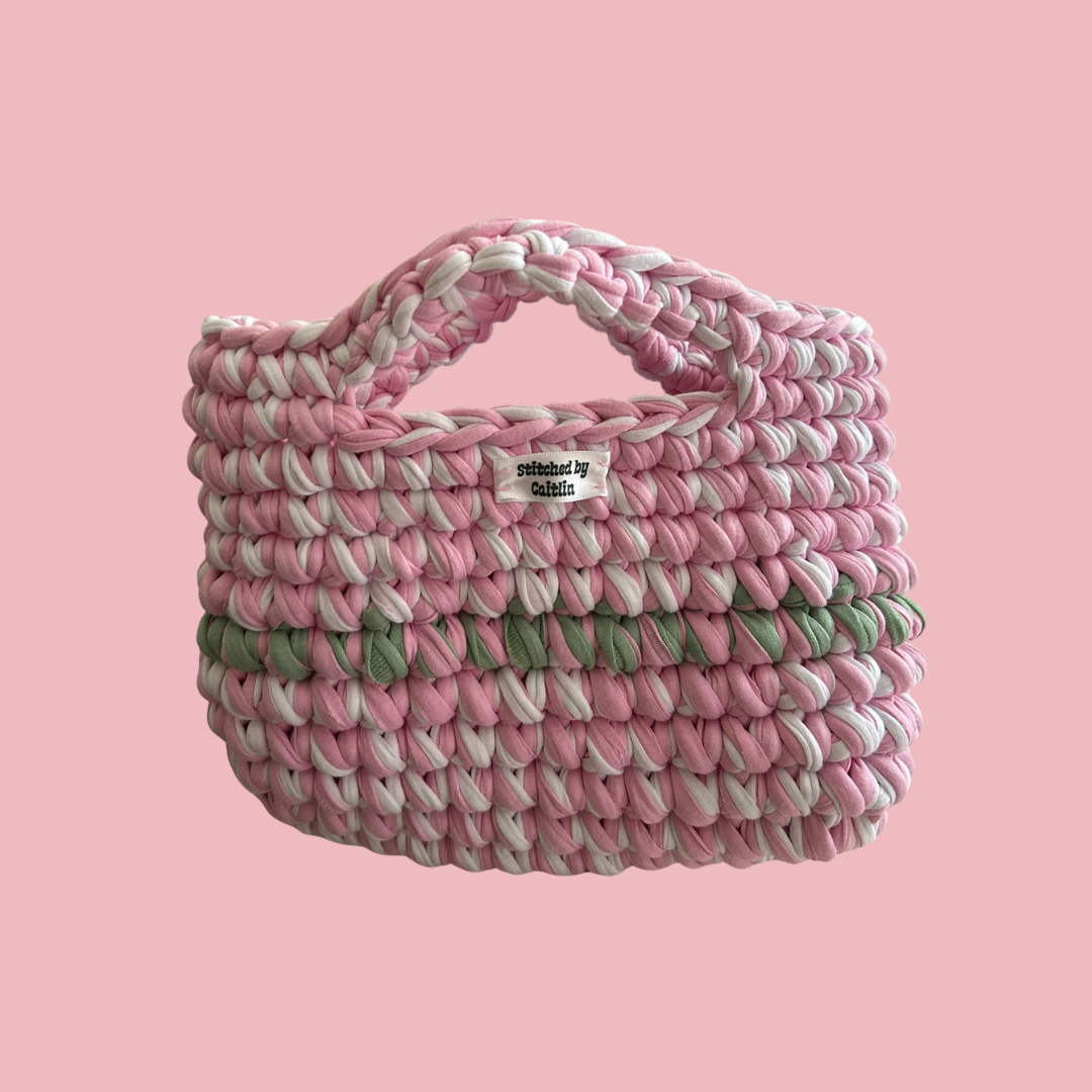 Pink, mint and white crocheted bag with two small handles at the top and a Stitched by Caitlin label on the front