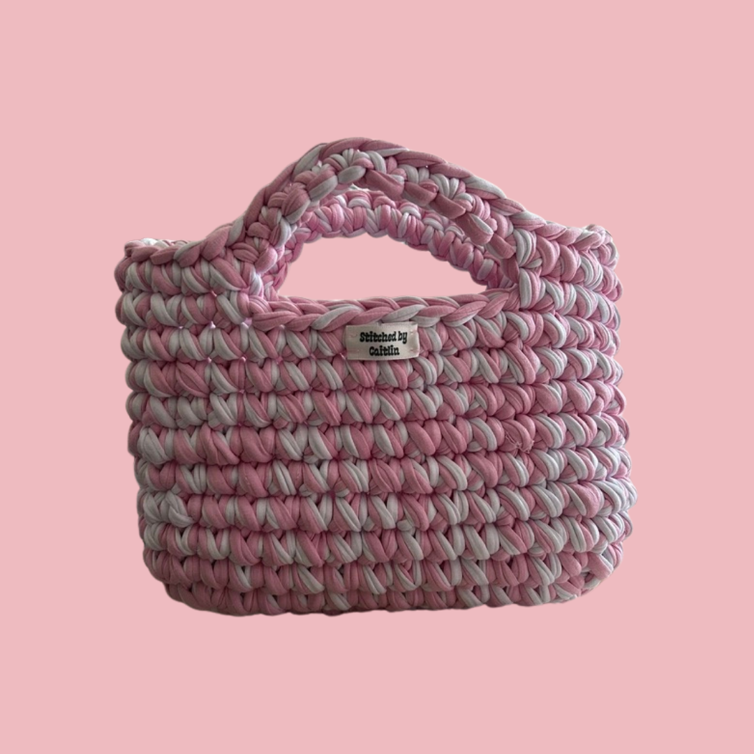Pink and white crocheted bag with two small handles at the top and a Stitched by Caitlin label on the front