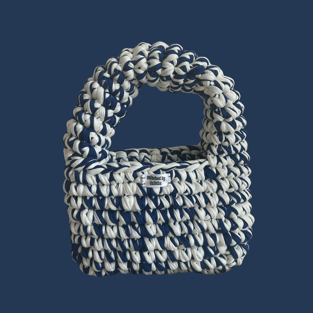 Blue and white crocheted bag with a half-moon bubble strap at the top and a Stitched by Caitlin label on the front