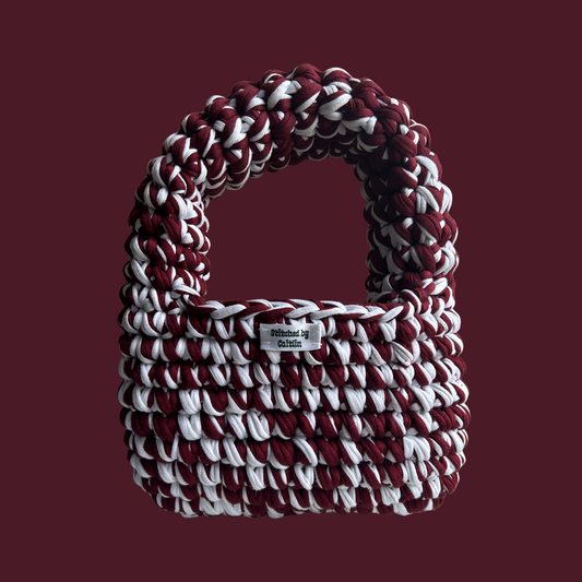 Burgundy and white crocheted bag with a half-moon bubble strap at the top and a Stitched by Caitlin label on the front