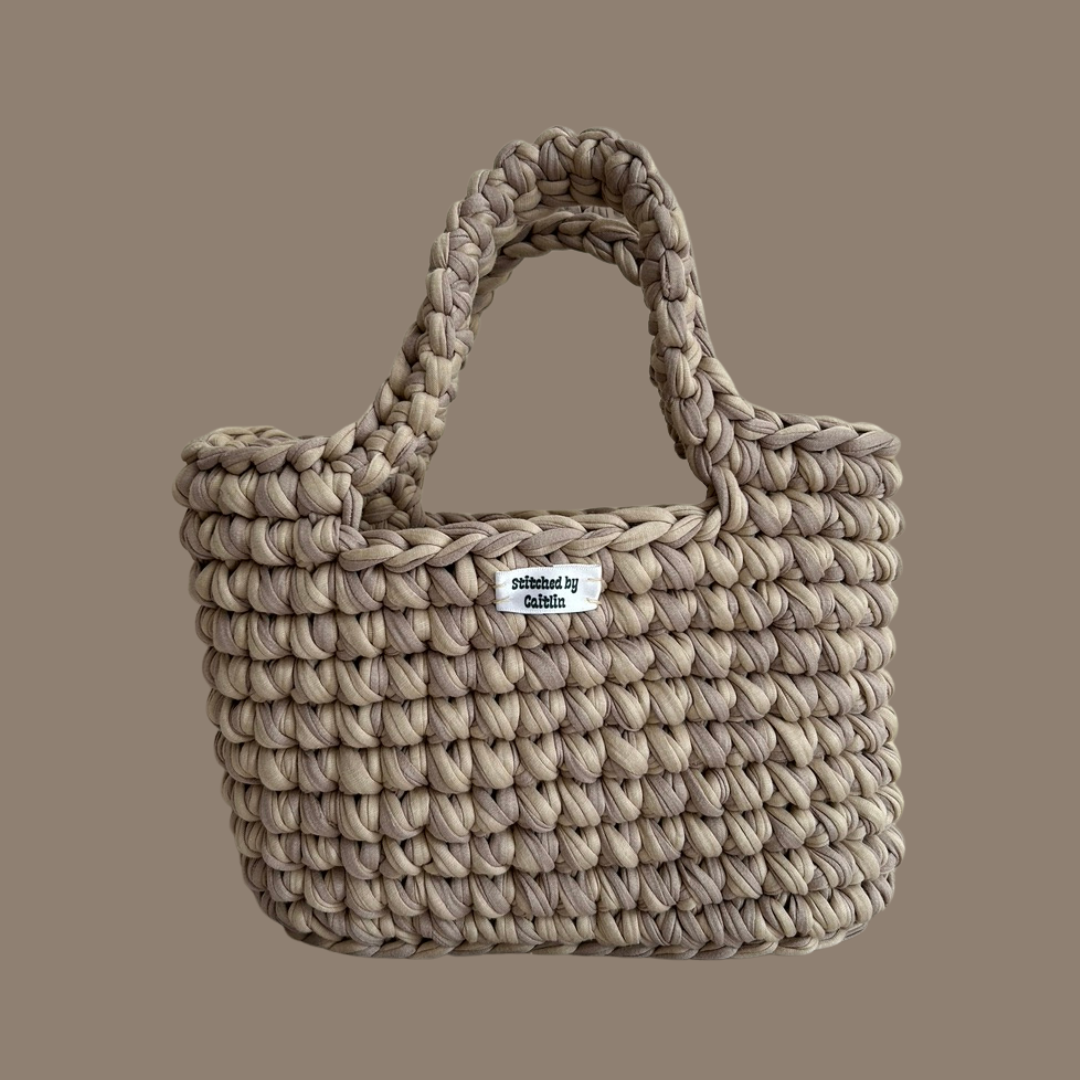 Light and medium brown crocheted bag with two long handles at the top and a Stitched by Caitlin label on the front
