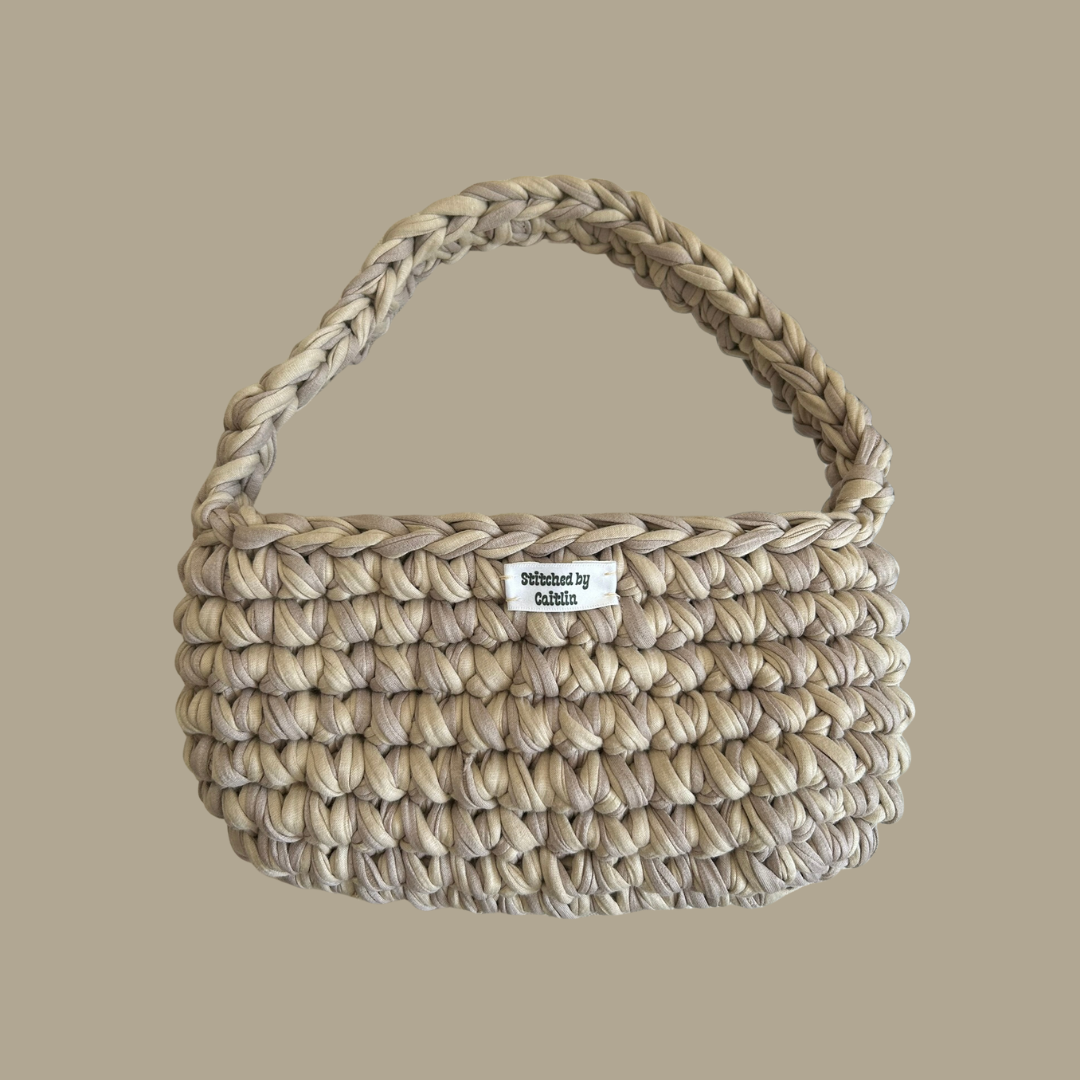 Biege and light brown crocheted bag with a single shoulder strap at the top and a Stitched by Caitlin label on the front