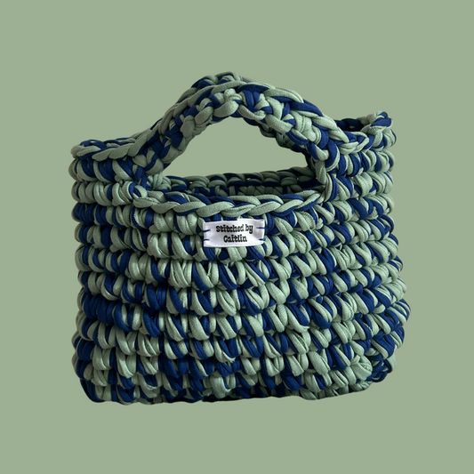 Mint green and blue crocheted bag with two small handles at the top and a Stitched by Caitlin label on the front