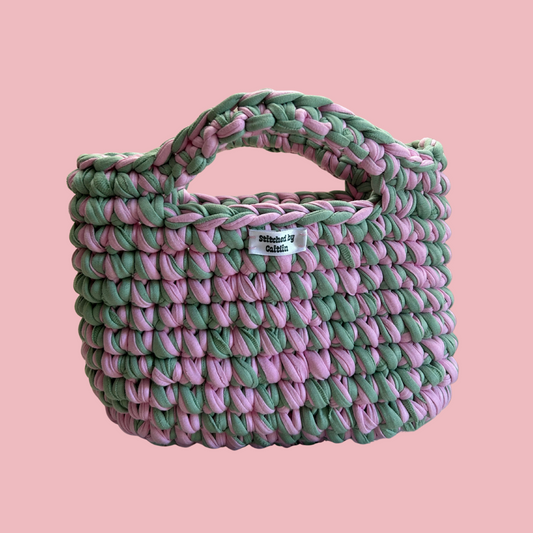 Pink and mint crocheted bag with two small handles at the top and a Stitched by Caitlin label on the front