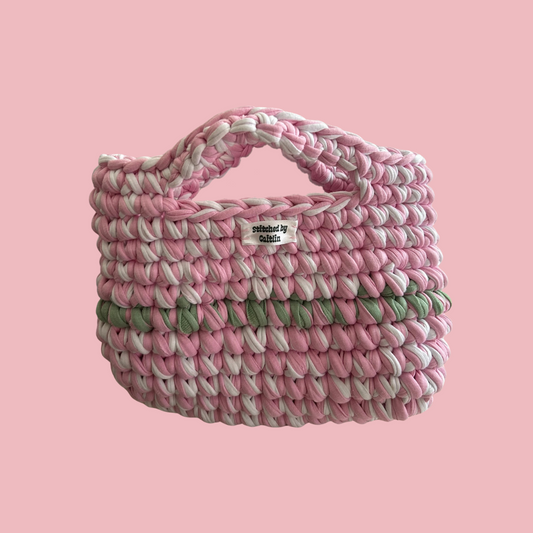 Pink, mint and white crocheted bag with two small handles at the top and a Stitched by Caitlin label on the front