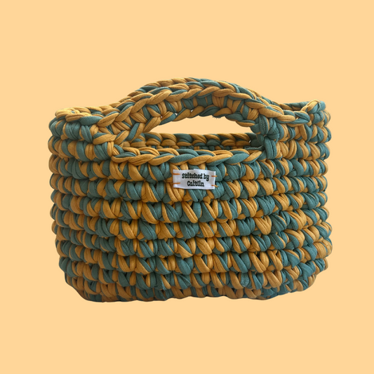 Yellow and turquoise crocheted bag with two small handles at the top and a Stitched by Caitlin label on the front